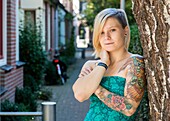 Tilburg, Netherlands. Young blonde woman with a tattoo on her left arm leaning against a tree in a domestic street.