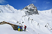 Three persons back-country skiing standing in front of alpine hut, Tete dell'Autaret and Pelvo di Ciabriera in background, Monte Faraut, Valle Varaita, Cottian Alps, Piedmont, Italy