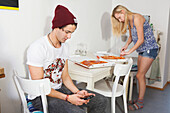 Young couple in the kitchen, playing with mobile and eating pizza