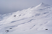 Drei kleine Holzhäuser im verschneiten Hochtals namens Fondei, dahinter ein namenloser Gipfel oberhalb der Strassberger Alp, Bündner Alpen, Kanton Graubünden, Schweiz
