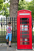 Teenage boy on mobile phone stands next to red telephone booth, London, England, United Kingdom
