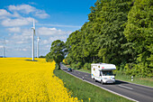 Motorhome camper on rural road and wind turbines in blooming canola field, near Alsfeld, Vogelsberg, Hesse, Germany