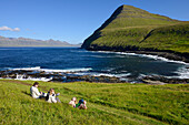 Familie sitzt in Wiese über der Bucht bei Gjogv, Insel Eysturoy, Färöer Inseln (Føroyar)