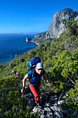 Eine junge Frau wandert durch Macchia an der Küste, Felsnadel bei Pedra Longa und Santa Maria Navarrese im Hintergrund, Selvaggio Blu, Sardinien, Italien, Europa