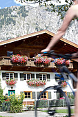 Young woman riding past a house on her bike on a sunny day, Tannheimer Tal, Tyrol, Austria