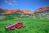 Woman sitting in tent and eating, Lechtal Alps in alpenglow in background, valley Fundaistal, Lechtal Alps, Tyrol, Austria