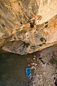 A women in shorts and a blue top rock climbing on a steep route above water in Rodeallr Spain. There is blue a raft at the base of the rock wall where the climber's had rafted in to access the cliff.