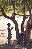 Young Himba boy standing in the shade of a tree in semi-silhouette, Kunene Region (formerly Kaokoland) in the far north of Namibia