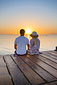Young couple sitting at the end of a jetty in the morning atmosphere and enjoying the view of the sunrise. Playa de Muro beach, Alcudia, Mallorca, Balearic Islands, Spain