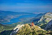 Frau beim Wandern sitzt auf Grasabsatz und blickt auf Lac d'Annecy, La Tournette, Hochsavoyen, Frankreich