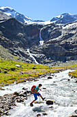 A male hiker jumping from stone to stone to cross a river, during the Glocknerrunde, a 7 stage trekking from Kaprun to Kals around the Grossglockner, the highest mountain of Austria.     Located in the heart of the Hohe Tauern National Park which contrast