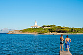 Girls Enjoying The View Of Alcanada Island And Its Lighthouse Near Alcudia, Mallorca, Balearic Islands, Spain