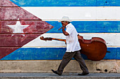 Hispanic musician carrying upright bass in front of Cuban flag mural