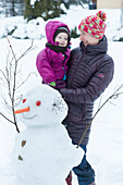 Mutter mit Tochter auf dem Arm vor Schneemann, 1 Jahr alt, Familie im Winter, Schnee, Holzhau, Sachsen, Deutschland