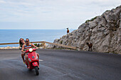 Young couple riding a red scooter on road along Cap de Formentor peninsula with goats on rocks, Palma, Mallorca, Balearic Islands, Spain