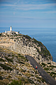 Young couple riding a red scooter on road along Cap de Formentor peninsula with Faro de Formentor lighthouse behind, Palma, Mallorca, Balearic Islands, Spain