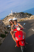 Young couple riding a red Vespa scooter on a coastal road along the Cap de Formentor peninsula with Faro de Formentor lighthouse behind, Palma, Mallorca, Balearic Islands, Spain