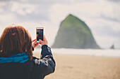A young woman uses her smartphone to take a picture of Haystack Rock while visiting Cannon Beach, Oregon.