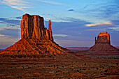 The East and West Mitten Buttes are illuminated by the reddish-yellow sun against a blue and purple sky at sunset in Monument Valley Navajo Park