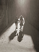 Two children running hand in hand down a sunlit alley, France, Europe