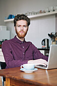 Portrait of confident young man using laptop at table in kitchen