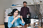 Portrait of young woman having coffee at kitchen table with man in background