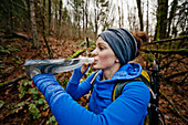 A young active woman pauses to take a drink while hiking through a forest.