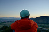 Young man in outdoor clothing and cap is sitting on rocks is looking on sunset in Macedonia.