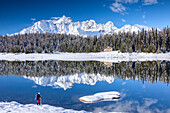 Hiker admires the snowy peaks and woods reflected in Lake Palu, Malenco Valley, Valtellina, Lombardy, Italy, Europe