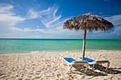 A palm leaf beach umbrella and two beach chairs on a sandy beach in Cayo Coco, Cuba.