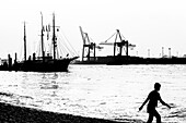 Silhouette einer Frau die in Oevelgönne vor der Kulisse des Museumshafens und dem Containerterminal Burchardkai am Strand spazieren geht, Hamburg, Deutschland