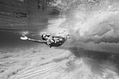 Black and white photo of a girl in a bikini swimming underwater in the ocean.  She goes underneath the waves over a sandy bottom covered by clear water.