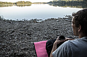 Young man sitting at a lake and drinking coffee, Freilassing, Bavaria, Germany