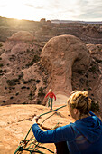 A woman lowering a young girl with a rope while rappelling down a sandstone prow at sunset, Kane Creek Road, Moab, Utah.
