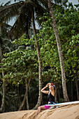 Junge Surferin sitzt am Strand und trinkt aus einer Kokosnuss, Sao Tome, Sao Tome und Príncipe, Afrika