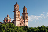 The itricate decorations of Taxco Cathedral's tower stand proud over the treeline, against a blue sky