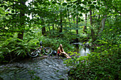 Junge Frau sitzt an Bachlauf im Wald mit Mountain Bike neben ihr, Kleinkahl, nahe Schöllkrippen, Kahlgrund, Spessart-Mainland, Bayern, Deutschland