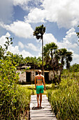 A young woman in board shorts and bikini top walks on a wooden dock toward an ancient ruin in a lush, green tropical scene.