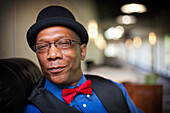 An african american man wearing a hat, vest and a red bow tie looks forward.