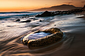 Water washes up onto the sand and a rock on the beach with an orange sky glowing at sunset, Tarifa, Cadiz, Andalusia, Spain
