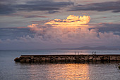 A stone pier out in the tranquil water reflecting the glowing clouds at sunset over the mediterranean, Lacco Ameno, Ischia, Naples, Campania, Italy