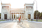 Young woman bicycling in front of the prpypaea on Königs Plaza in Munich, Bavaria, Germany