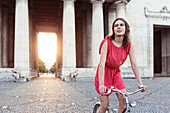 Young woman bicycling in the evening sun through the propylaea, Königs Plaza in Munich, Bavaria, Germany