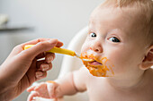 Mother feeding messy baby son with spoon in high chair