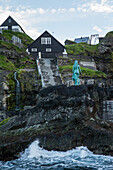 Statue einer Frau auf einem Felsen im Meer vor einem Dorf, Färöer Inseln