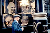 Man walking along the street with cap and jacket, a lamp post in the foreground and several portraits of famous people in background. London, England