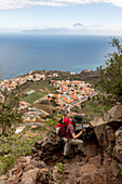 Wanderin mit rotem Rucksack, Blick Wanderweg zum Mirador de Abrante, Steilwand, in der Ferne der Pico del Teide, Teyde, höchste Berg Spaniens, Insel Teneriffa, unten liegt Agulo auf La Gomera, Kanaren, Kanarische , Inseln, Spanien
