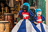 Two rag dolls, one black and one white, dressed in Cuban flag dresses, hanging from the gate of a souvenir shop. Behind them the shop is full of handcrafted drums and musical instruments. Old Havana (Havana Vieja), La Habana, Cuba