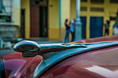 'Close up of a vintage Dodge ram'' hood ornament on a burgundy red vintage car. People are out on the sidewalk of busy Avenida Galiano in the background. Centro Havana, La Habana, Cuba'''