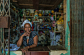A proud Cuban woman leans on the counter of her Botanica stall, displaying shelves of vases, masks, bracelets and beaded necklaces, and other tools of the trade. She wears a star-patterned shirt and a headscarf. Old Havana (Havana Vieja), La Habana, Cuba